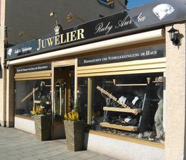Außenansicht eines Juweliergeschäfts mit Schmuck-Auslagen und Werbeschildern im Fenster.
