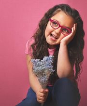 Niña con gafas rosadas sonríe y sostiene flores en fondo rosa.