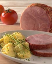 Gekochter Schinken mit Kartoffelsalat und Schnittlauch auf einem Teller, zwei Tomaten im Hintergrund.