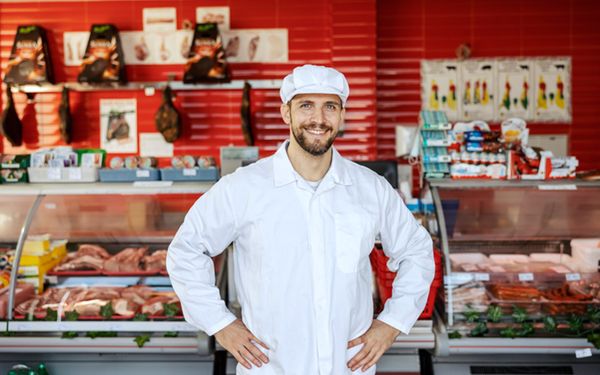 Metzger in weißer Kleidung steht vor einer Metzgereitheke mit Wurst und Fleisch