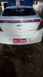 Táxi Chevrolet Prisma branco estacionado, placa vermelha, em piso de blocos.