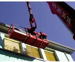 Roter Kran hebt Arbeitsplattform vor einem Gebäude hoch, Person hinter gelbem Fensterrollo sichtbar.