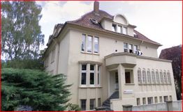 Großes weißes Gebäude mit roten Dachziegeln und mehreren Fenstern, umgeben von Bäumen.