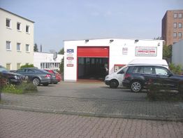 Autos parken vor einer Werkstatt mit rotem Tor in städtischer Umgebung bei klarem Himmel.