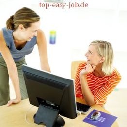 Zwei Frauen lächeln sich an, eine sitzt vor einem Computer, die andere steht daneben.