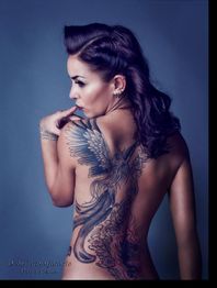 Frau mit großem Phönix-Tattoo auf dem Rücken, mit seitlichem Haarstil, posiert vor blauem Hintergrund.