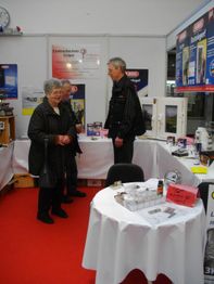 Eine ältere Frau und ein Mann stehen an einem Messestand mit Prospekten und Ausstellungsstücken.