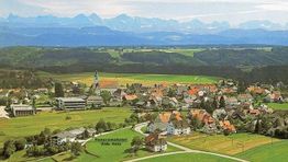 Luftaufnahme eines Dorfes mit Kirche, Feldern und Bergen im Hintergrund.