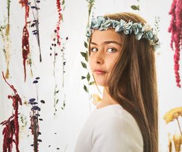 Niña con corona de flores azules, rodeada de ramas y plantas colgantes en fondo claro.