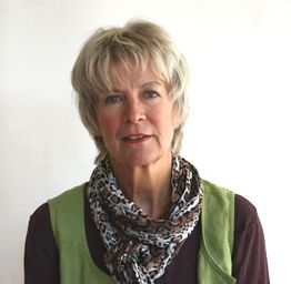 Eine Frau mit kurzem blondem Haar und Schal vor weißem Hintergrund, trägt grüne Weste.