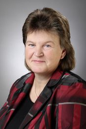Frau mit kurzen braunen Haaren und rot-schwarzem Blazer vor grauem Hintergrund.