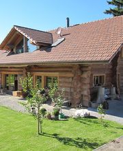Blockhaus mit rotem Dach, Garten und kleinem Baum im Vordergrund. Sonniger Tag, blauer Himmel.