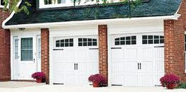 Two white garage doors with windows, flanked by red brick walls and potted flowers in front.