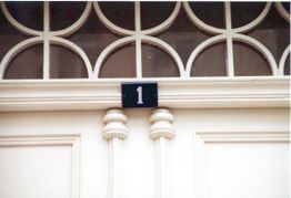 Hellgraue Tür mit Nummernschild "1", verziert mit runden Glasfenstern darüber.