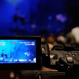 Close-up of a video camera recording a dimly lit, blue-toned stage performance.