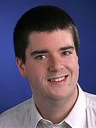 Ein Mann in einem weißen Hemd vor blauem Hintergrund lächelt in die Kamera.