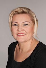 Eine lächelnde Frau mit blondem Haar in schwarzem Oberteil vor grauem Hintergrund.