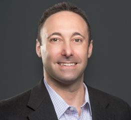 Man smiling, wearing a dark suit jacket and light shirt, on a neutral gray background.