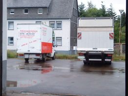 Zwei geparkte LKWs auf einem nassen Hof vor einem grauen Gebäude.