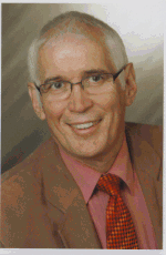 Älterer Mann im Anzug mit Brille lächelt in die Kamera vor neutralem Hintergrund.