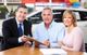 A man with a clipboard helps a couple signing papers at a car dealership table.