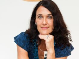 Eine Frau mit dunklem Haar lächelt in die Kamera, trägt ein blau gemustertes Oberteil.