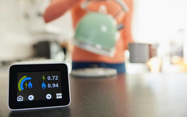 Ein Smart Meter steht auf einer Küchenarbeitsfläche und zeigt den Verbrauch im Haushalt an. Im Hintergrund kocht sich eine Frau im orangenen Oberteil einen Tee.