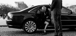 Une femme sort d'une voiture de luxe noire, accueillie par un homme en costume.