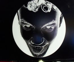 Rostro en negativo, cabello rizado, dentro de un círculo blanco sobre fondo negro.