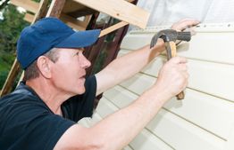 Man in blue cap nailing siding to a house wall.