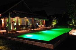 Haus mit beleuchtetem Pool im Garten bei Nacht, umgeben von grüner Beleuchtung.