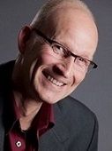 Eine lächelnde Person mit Brille in Anzug und Hemd, seitlich fotografiert vor grauem Hintergrund.