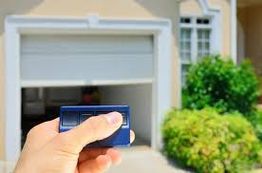 Hand holding a remote control in front of an open garage door with greenery nearby.