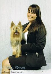 Eine Frau im Anzug sitzt mit einem fröhlichen Australian Silky Terrier auf dem Schoß vor hellem Hintergrund.