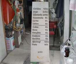 Steinsäule in Werkstatt mit Städtenamen und Jahreszahl 1723, umgeben von Werkstattutensilien.