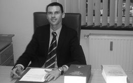 Ein Mann in einem Anzug sitzt an einem Schreibtisch und schreibt. Bücher liegen vor ihm.