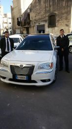 Due uomini in giacca e cravatta accanto a un'auto bianca parcheggiata in una strada cittadina.