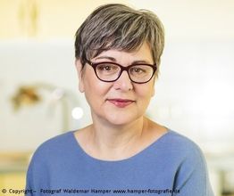 Eine Person mit grauem Haar und Brille, trägt einen blauen Pullover und lächelt in die Kamera.