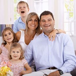 Smiling family of five sitting together in a bright room, with two girls, a boy, and parents.
