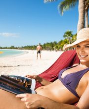 Mujer sonriente lee revista en la playa, con sombrero y bikini, bajo el sol y palmeras.