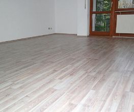 Leerer Raum mit hellem Laminatboden, weißer Wand und großem Fenster mit Blick ins Grüne.