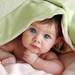 Baby with blue eyes under a green blanket, looking up with a curious expression.