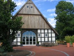 Fachwerkhaus mit Holzgiebel und Terrasse unter blauem Himmel, umgeben von Bäumen.