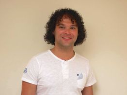 Persoon met donker krullend haar in wit T-shirt glimlacht voor een beige achtergrond.