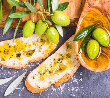 Pane con olio d'oliva, olive verdi e un cucchiaio di legno su un tavolo grigio.