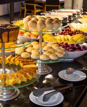Buffet de café da manhã com pães, bolos e frutas em mesas de vidro.