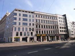 Mehrstöckiges Gebäude mit Geschäften im Erdgeschoss, Straße im Vordergrund, klarer Himmel oben.