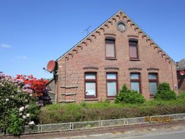 Backsteinhaus mit Satteldach, roten Fensterrahmen und Sat-Schüsseln, umgeben von Sträuchern.