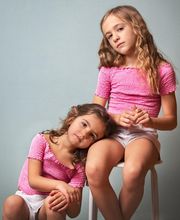 Dos niñas con blusas rosas, una sentada en un taburete y la otra apoyada en su rodilla.