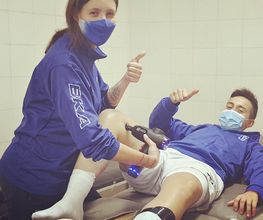 Persona aplica terapia en la pierna de un hombre, ambos con mascarillas y ropa azul.
